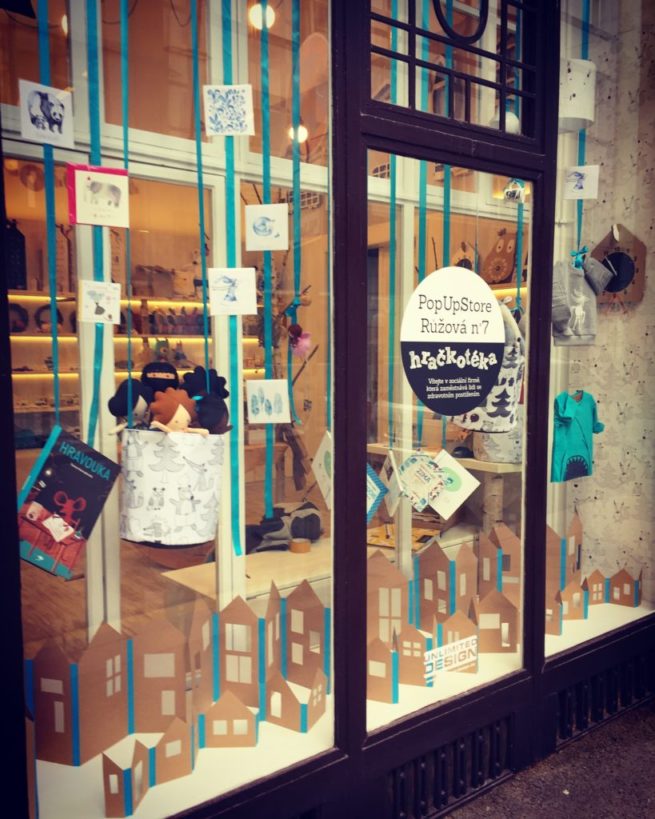 Výloha pro druhou pobočku hračkářství Hračkotéka v Praze. Design výkladní skříně, dekorace s barevnými stuhami a papírovým podsvětleným městem.